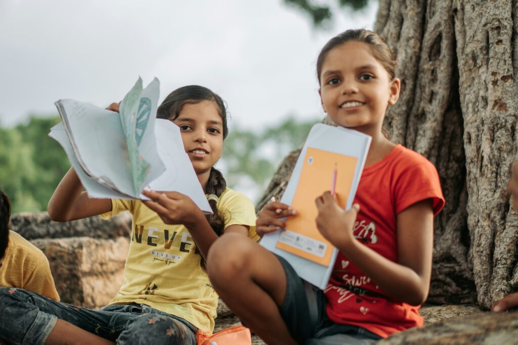 Two Indian girls smiling and holding notebooks while studying outdoors.